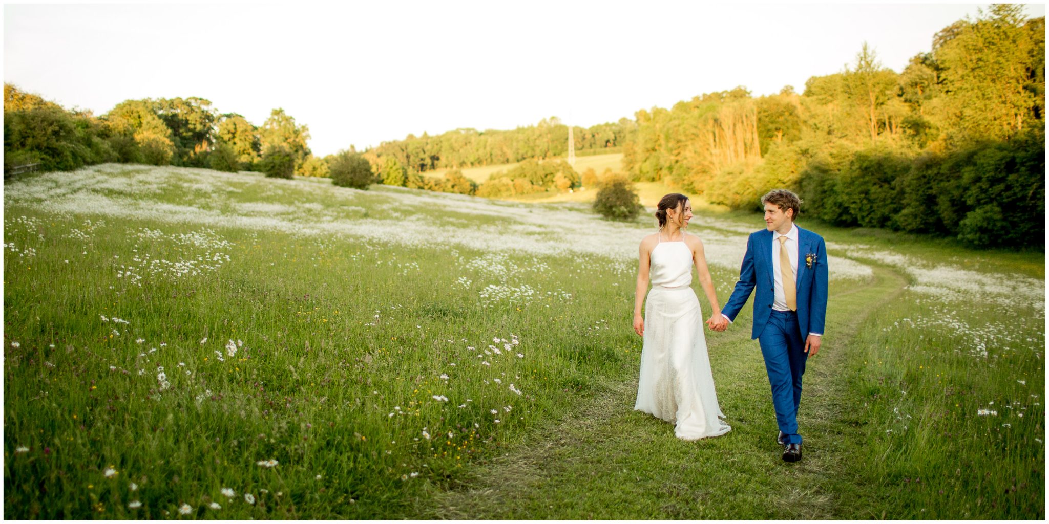 Bride and groom walk hand in hand through the meadow in the evening light