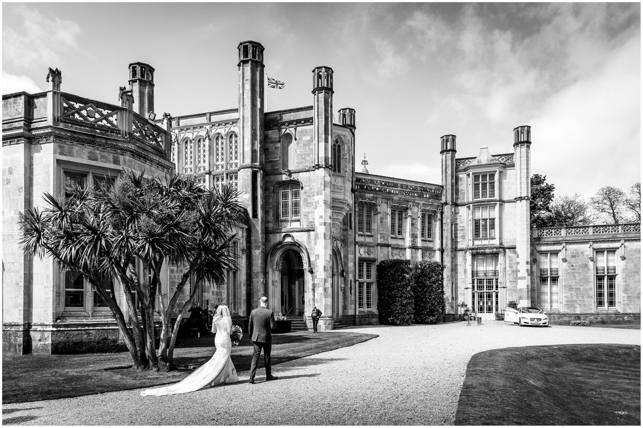 Black and white image of Highcliffe Castle with bride and groom walking towards wedding car by the main entrance